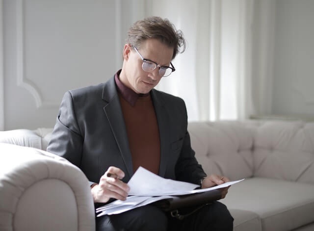 A man wearing a grey suit jacket and maroon sweater looking at a stack of papers