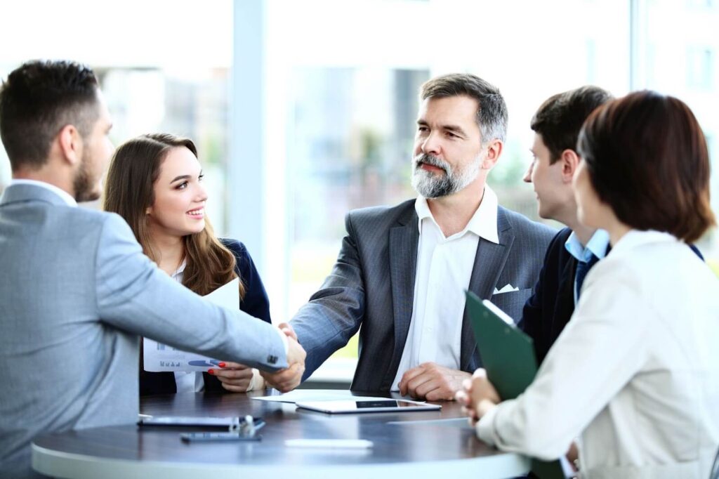 Man with grey hair and beard in a business suit shaking a younger man’s hand, while they are surrounded by colleagues.