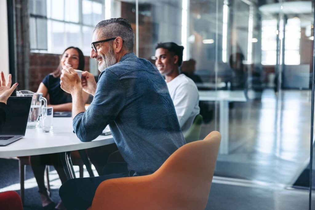 Man with grey hair, beard and glasses, drinking from a mug at his desk, surrounded by co-workers.